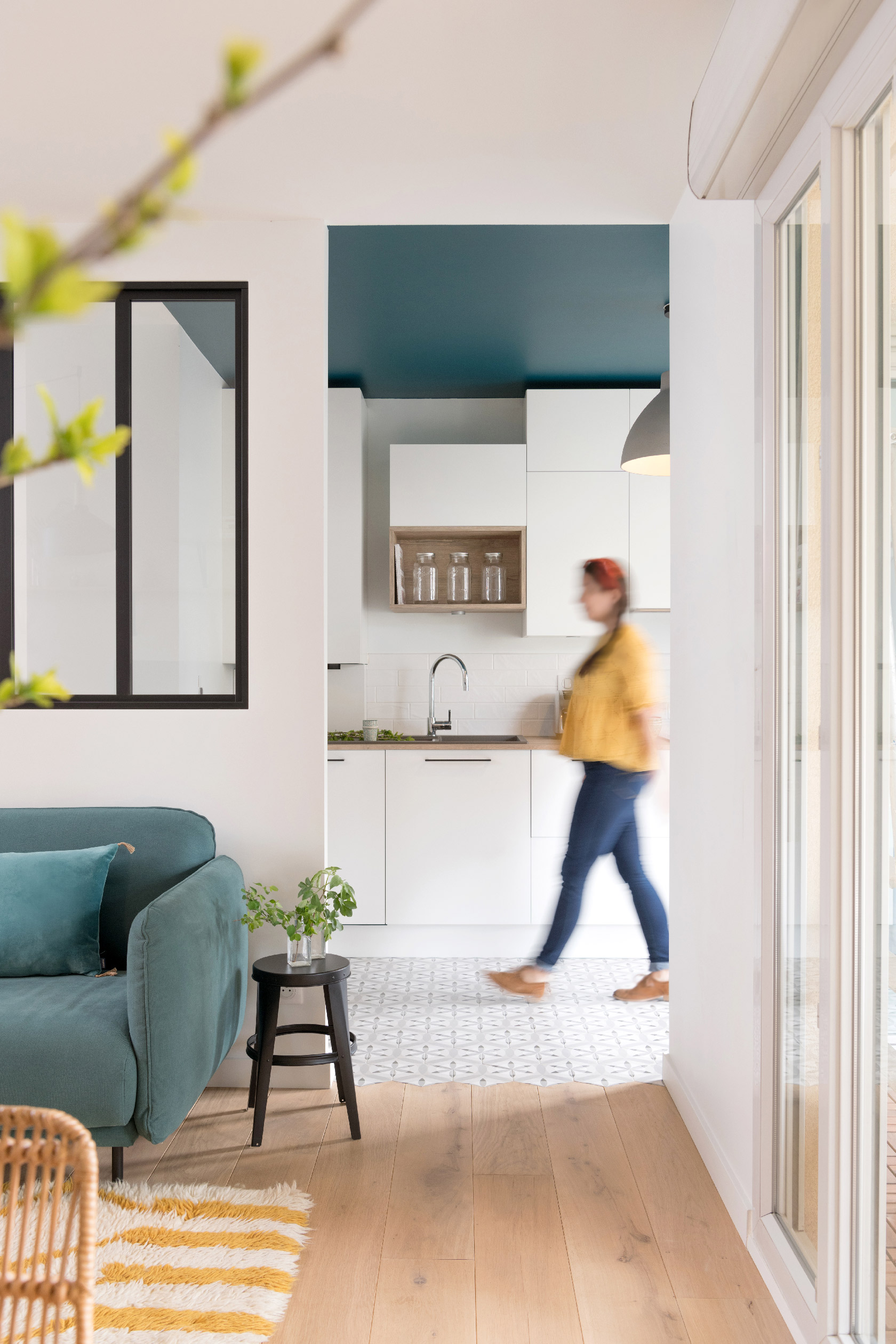 Intérieur d'un appartement moderne avec un coin cuisine, un canapé vert et un tabouret noir. Une personne passe devant, ajoutant une touche de dynamisme à l'espace.